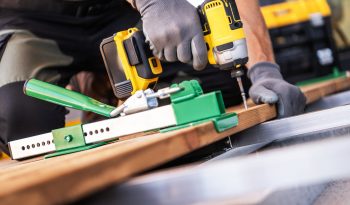 A carpenter is using a deckboard wrench and power drill to secure wooden boards on a construction site patio floor.